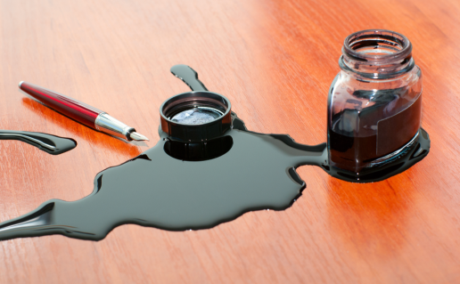 Spilled ink on wooden table next to half-full ink jar and fountain pen